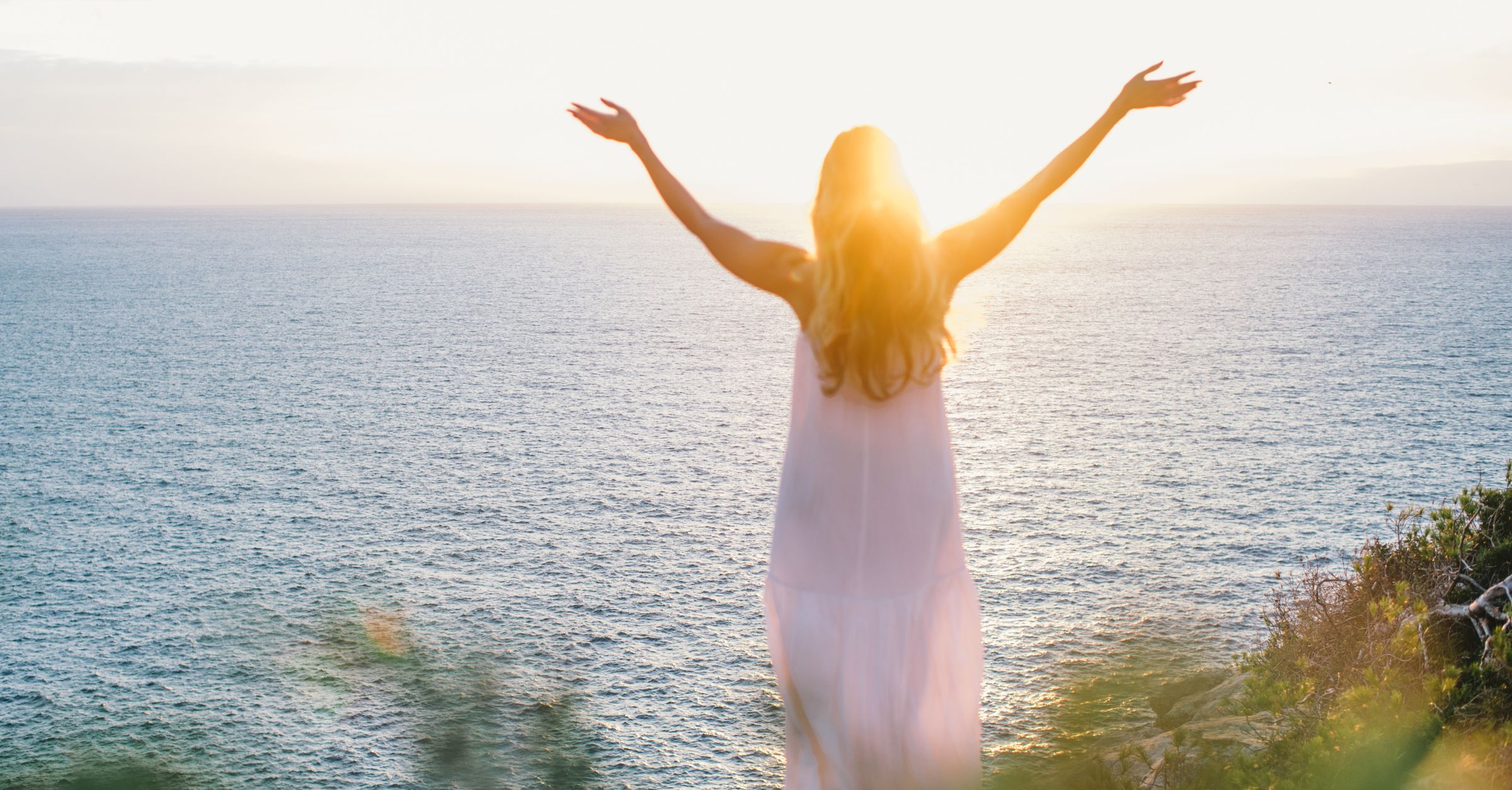 A woman looks off into the sunset with her arms raised, celebrating joy and success.