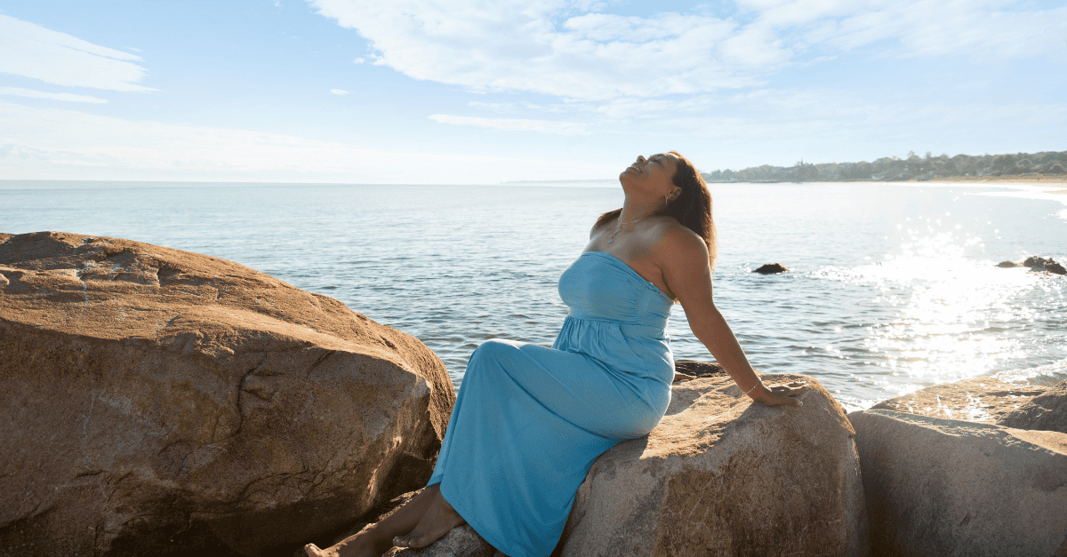 Beautiful woman in a blue dress takes a breath of fresh air while soaking up the sun on a boulder on the beach.